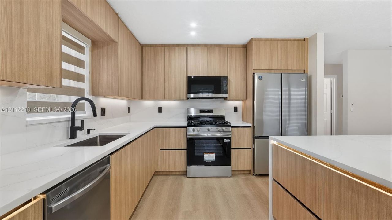 Kitchen featuring light wood-finish cabinetry, solid-surface countertops, full-height backsplash, stainless steel appliances, and matte black faucet