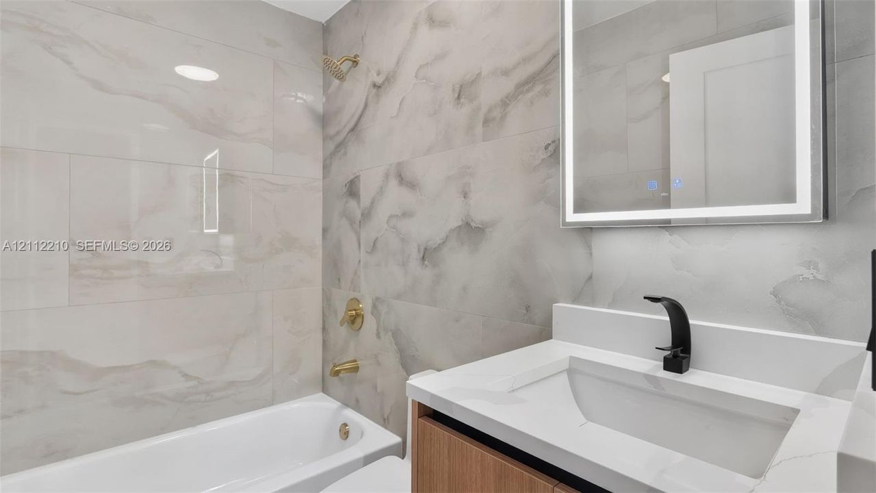 Bathroom featuring large-format stone-pattern tile surround, wood-finish vanity with white countertop, integrated basin, matte black faucet, and LED-backlit mirror