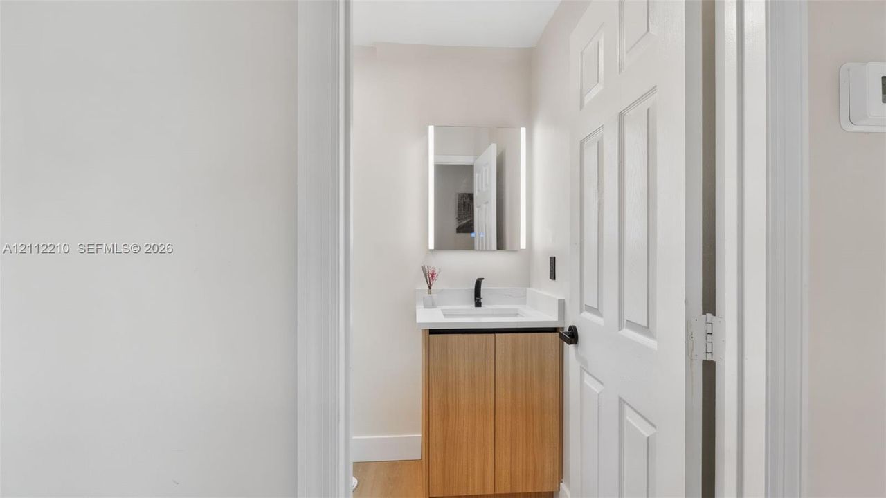 Light-toned wood vanity with a white countertop, integrated rectangular sink, matte black faucet, and rectangular LED-backlit mirror
