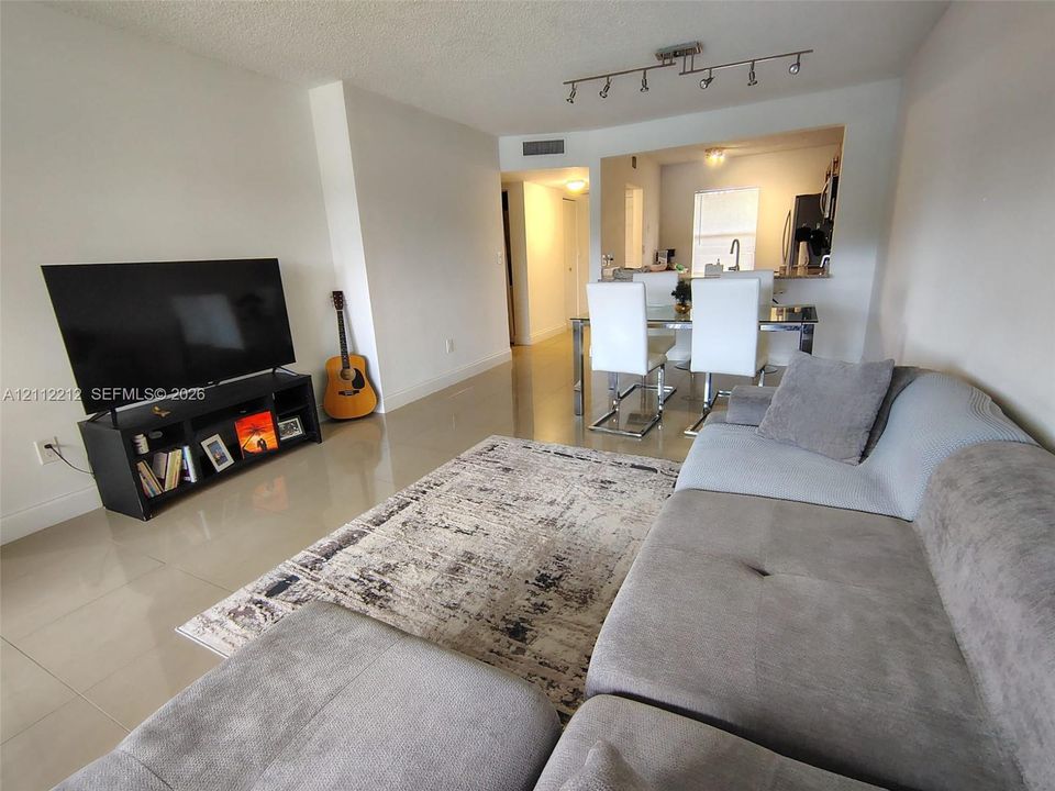 Open-concept interior featuring large-format tile flooring, recessed track lighting, and a kitchen pass-through window with an integrated breakfast bar