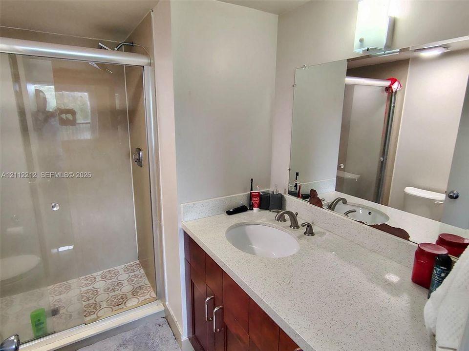 Bathroom featuring a vanity with wood-finish cabinetry, quartz countertop, and undermount sink, complemented by a glass-enclosed shower with patterned tile flooring and a wall-mounted mirror