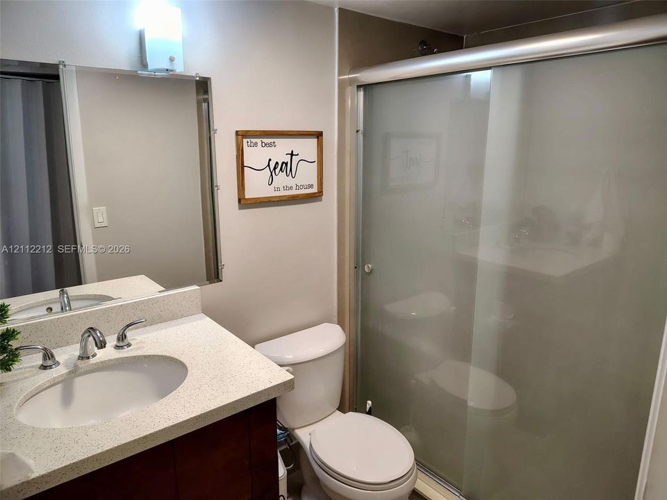 Bathroom featuring a vanity with a light-toned solid surface countertop, an undermount sink with dual-handle metal faucet, a rectangular wall-mounted mirror, and a walk-in shower with frosted sliding glass doors