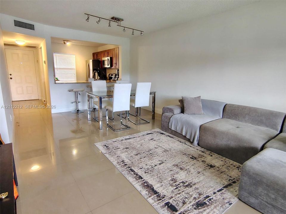 Open-concept interior featuring large-format tile flooring, recessed track lighting, and a kitchen pass-through window with a stone-surfaced breakfast bar
