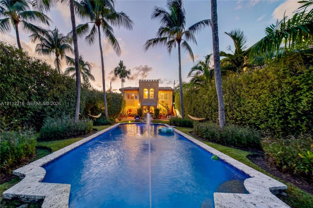 Rectangular in-ground pool featuring a central water fountain, stone-style pool coping, mature palm trees, and dense perimeter landscaping