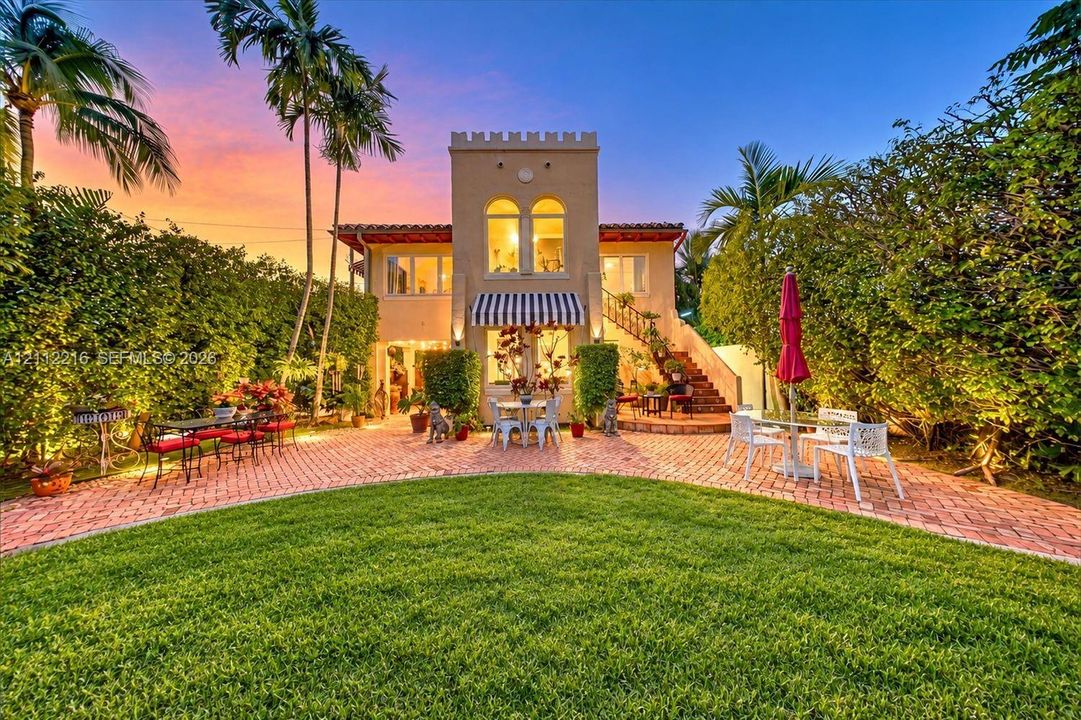 Mediterranean-inspired exterior featuring a crenelated roofline, brick paver patio, arched window detailing, exterior masonry staircase, and a manicured lawn