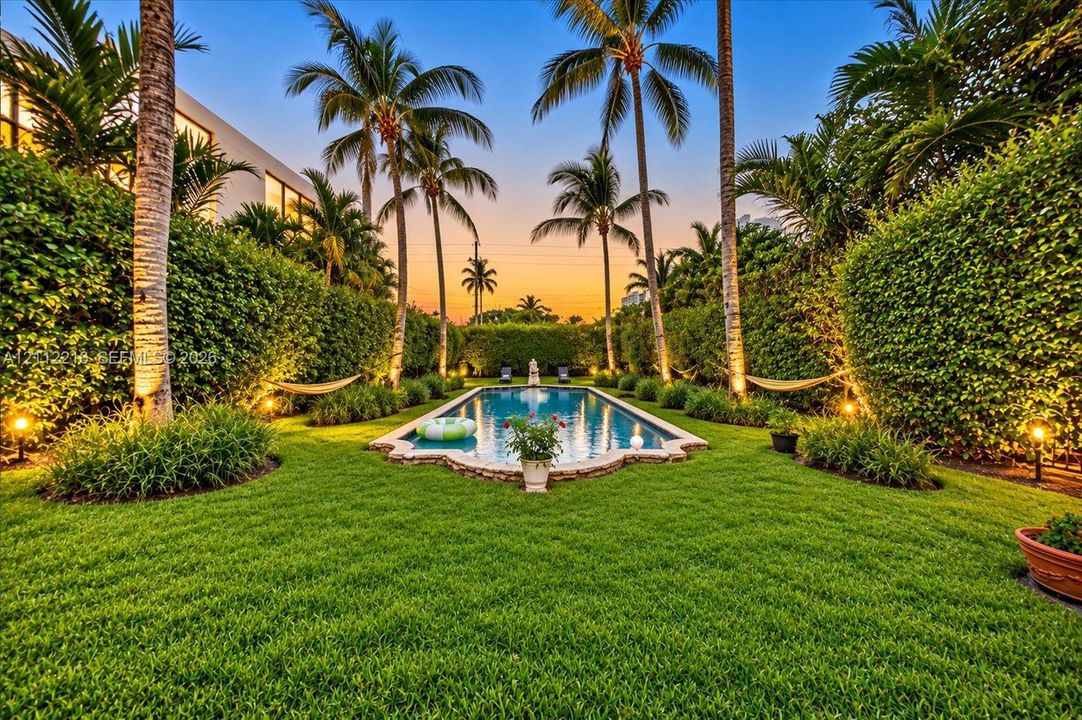 Rectangular swimming pool with stone coping, surrounded by a manicured lawn, established palm trees, and dense perimeter hedging