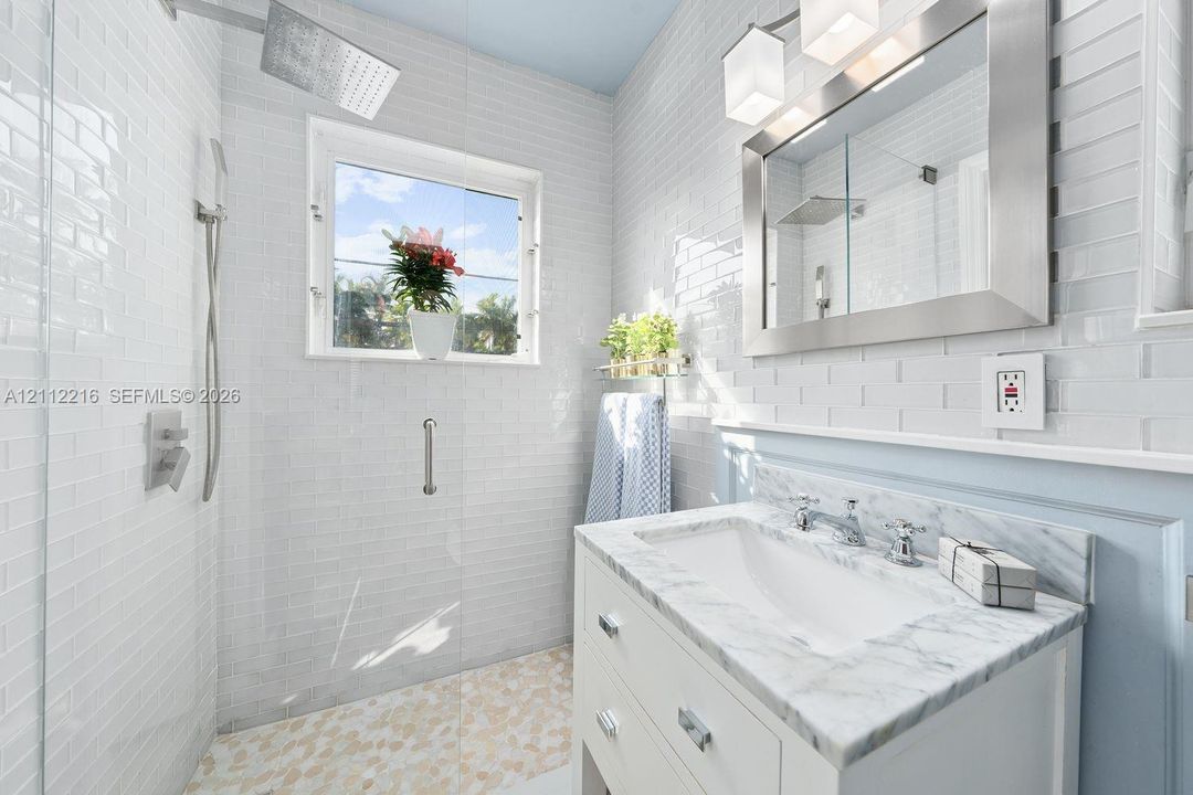 Bathroom featuring a white vanity with a stone countertop, a glass-enclosed shower with pebble-stone flooring, subway tile wall cladding, and a framed mirror with integrated lighting