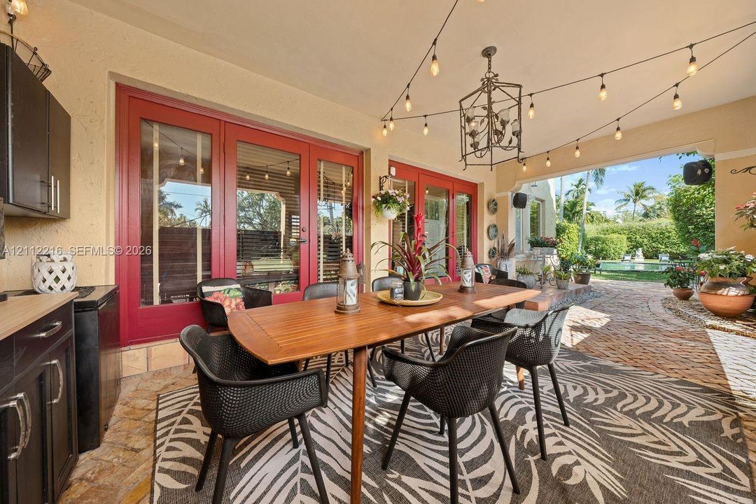 Covered patio featuring stone paver flooring, outdoor kitchen cabinetry, red-framed glass double doors, an overhead iron chandelier, and string lighting
