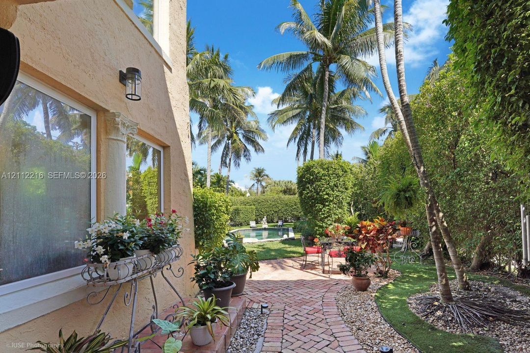 Stucco exterior with decorative column detail, brick-paved patio, lush landscaping with mature palm trees, and swimming pool