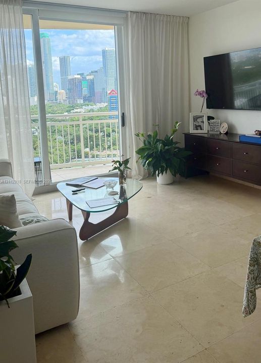 Floor-to-ceiling sliding glass doors opening to a balcony with metal railings and expansive city skyline scenery, complemented by large-format light-toned stone flooring