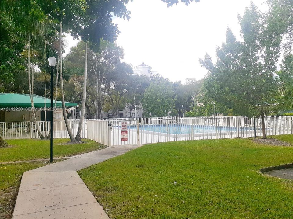 Swimming pool enclosed by white metal fencing, featuring an integrated cabana with a green canopy, concrete walkway, and mature landscaping