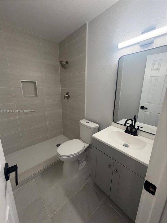 Bathroom featuring a walk-in shower with light-toned horizontal tile, a built-in recessed niche, a grey vanity with a white countertop, a rectangular mirror with a black frame, and matte black fixtures