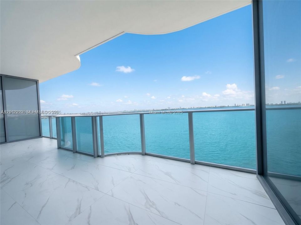 Expansive private balcony featuring large-format stone-patterned tile flooring, a curved architectural overhang, floor-to-ceiling glass panel railings, and full-height sliding glass doors