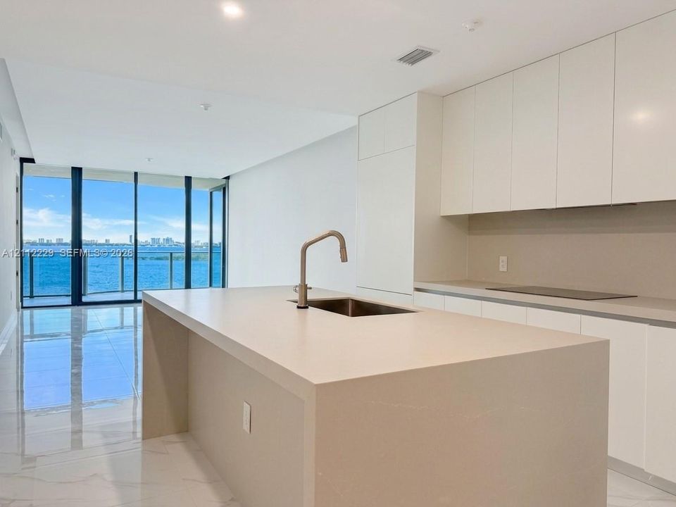 Modern kitchen featuring a central island with a waterfall countertop, integrated stainless steel sink, polished stone-finish flooring, floor-to-ceiling glass sliding doors, and minimalist cabinetry