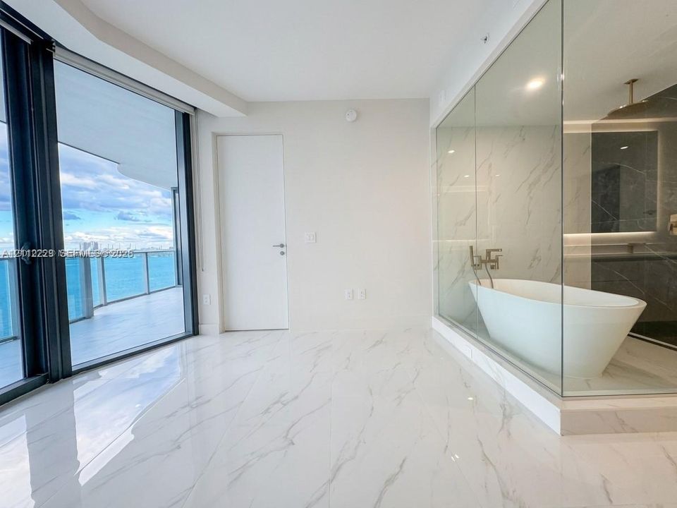 Floor-to-ceiling glass balcony doors, polished stone-finish flooring, glass-enclosed bathroom partition, freestanding soaking tub, and wall-mounted plumbing fixtures