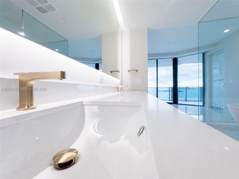 Minimalist vanity featuring a seamless white solid-surface countertop, integrated basin, brushed gold wall-mounted faucets, and an illuminated mirror