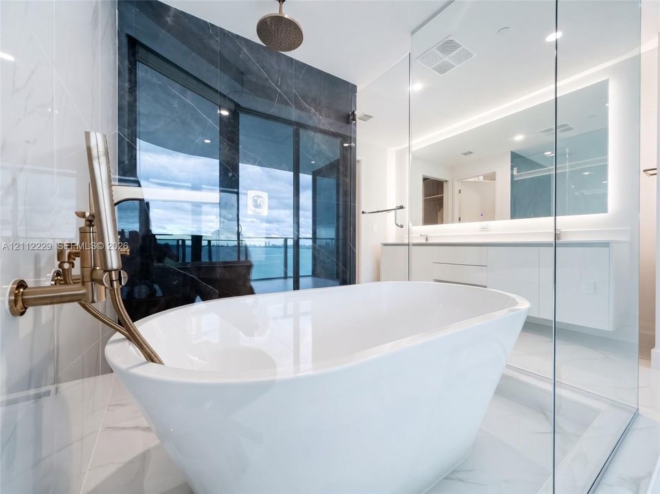 Freestanding soaking tub with brushed gold hardware, glass-enclosed shower, dual-vanity cabinetry, backlit wall mirror, and large-format tile flooring