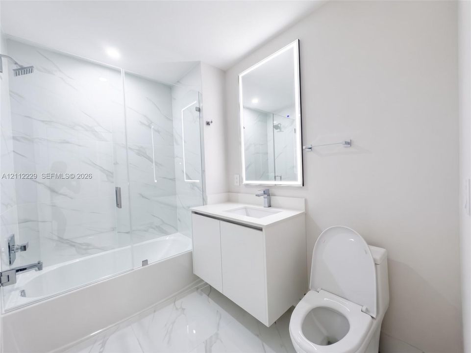 Bathroom featuring a glass-enclosed tub with floor-to-ceiling stone-style tile surround, floating vanity with a solid-surface countertop, and an integrated LED-lighted mirror