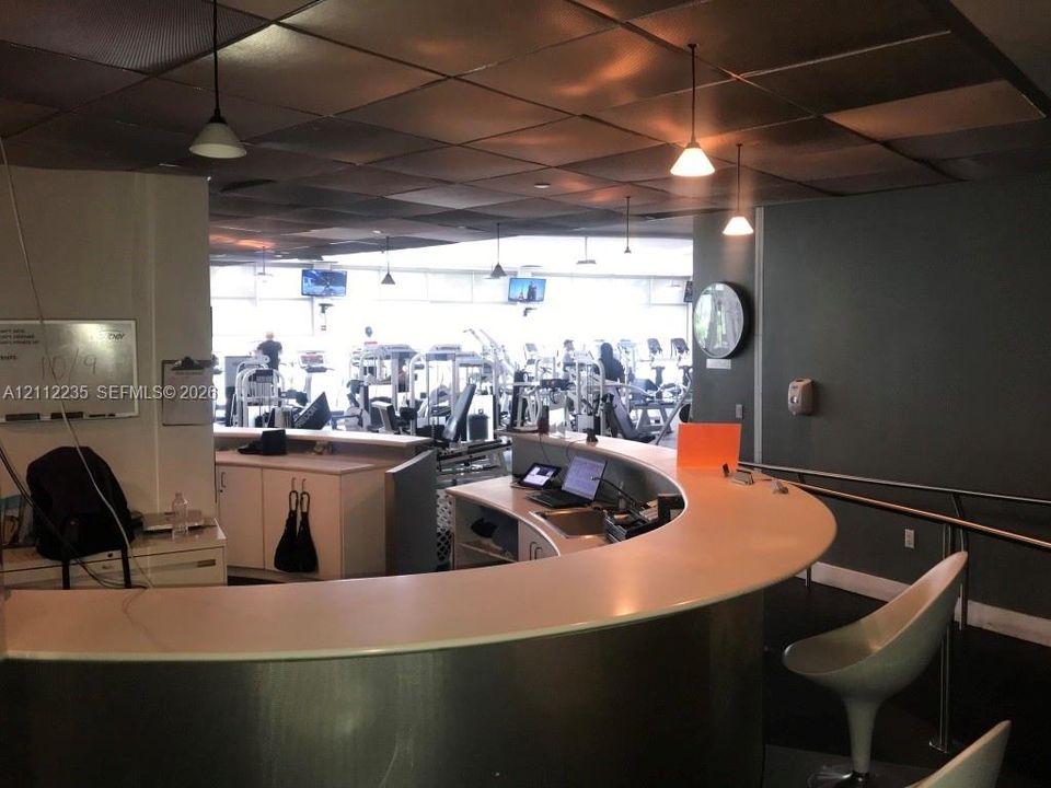 Curved reception desk with a smooth solid-surface countertop and metallic-finish base, drop-panel ceiling, and overhead pendant lighting