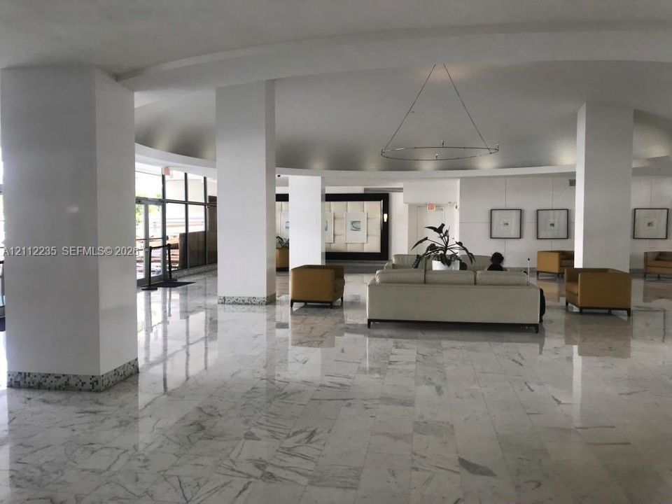 Spacious lobby featuring polished stone flooring, circular recessed ceiling detail, structural support pillars with speckled trim, and floor-to-ceiling glass paneling