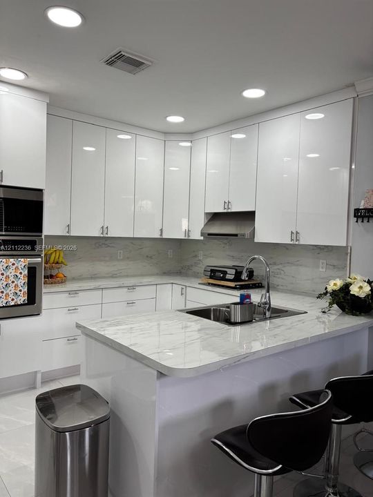 Kitchen featuring high-gloss white cabinetry, stone-patterned countertops, integrated stainless steel double oven, stainless steel range hood, and a double-basin undermount sink