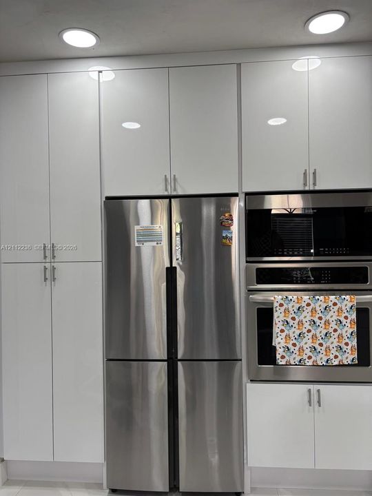 Floor-to-ceiling white high-gloss cabinetry, stainless steel French-door refrigerator, built-in stainless steel microwave, integrated stainless steel oven, and recessed ceiling lighting