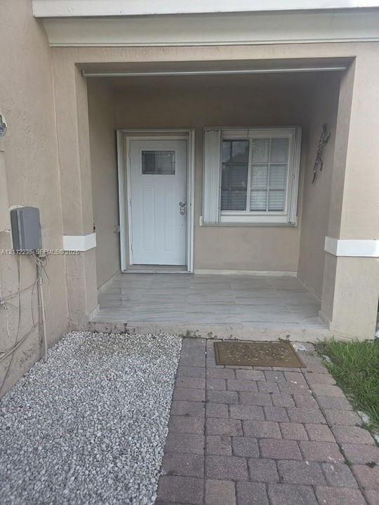 Covered entryway featuring a stone-tile porch, a white entry door with an integrated window, a sliding window with shutters, and a paver stone walkway