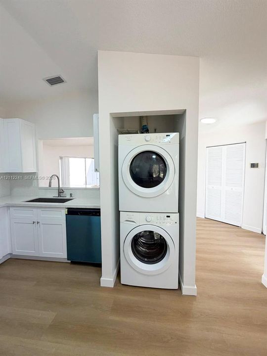 Stacked laundry unit featuring white appliances, recessed utility alcove, white cabinetry, solid-surface countertops, and wood-finish flooring