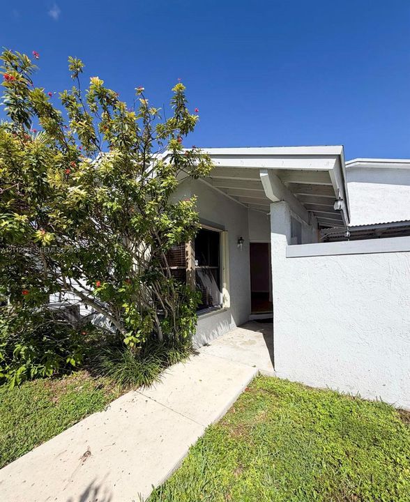 Stucco exterior featuring a covered entryway, exposed rafter tails, concrete walkway, and landscaped foliage