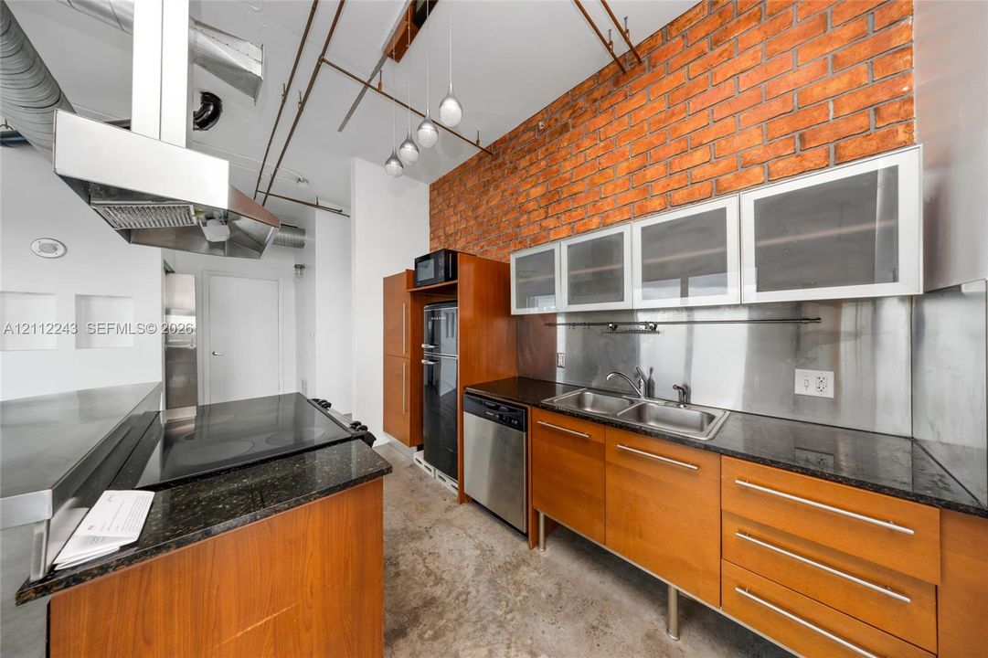 Kitchen featuring an exposed brick accent wall, stone-finish countertops, wood-finish cabinetry, stainless steel backsplash, and built-in stainless steel appliances