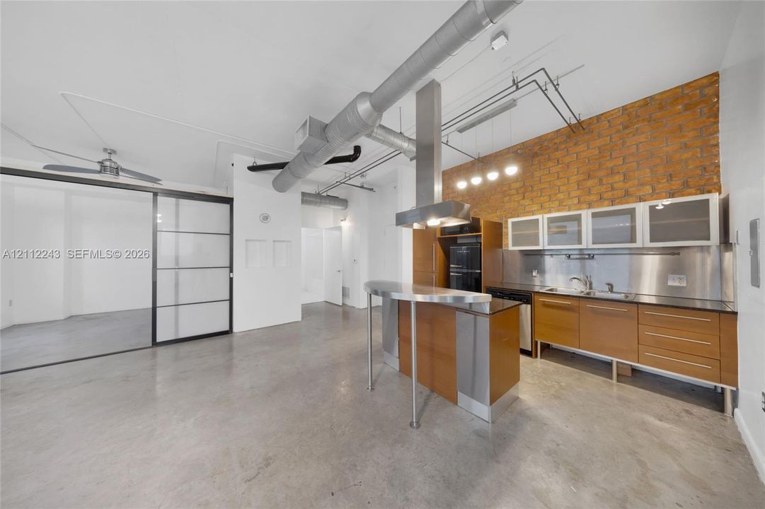 Industrial-style interior featuring exposed brick walls, polished concrete flooring, stainless steel backsplash, custom cabinetry with glass-panel upper storage, and stainless steel kitchen peninsula with integrated venting hood