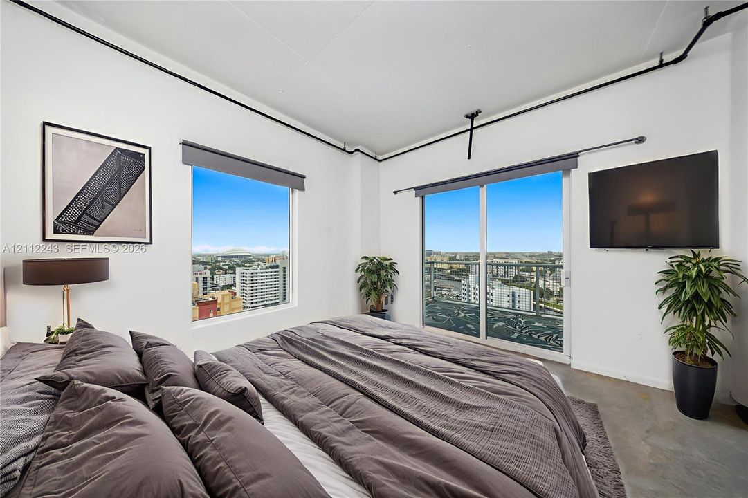 Industrial-style room featuring polished concrete flooring, exposed ceiling-mounted piping, sliding glass door to a private balcony, and large windows with urban exterior scenery