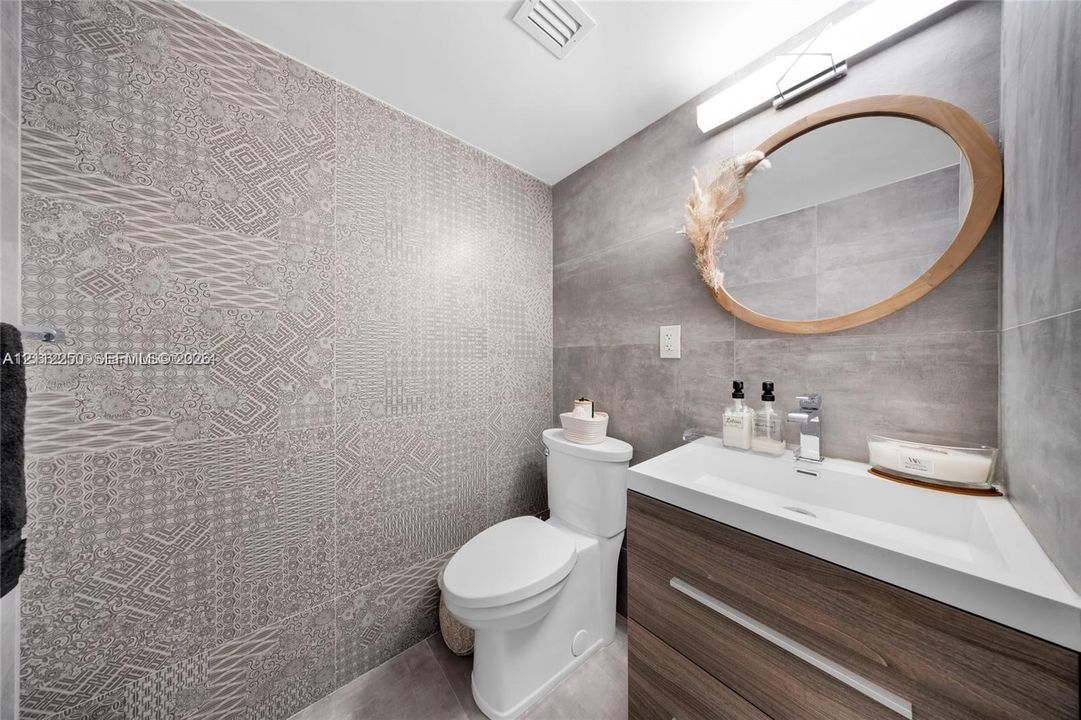 Powder room featuring a wood-finish vanity with an integrated basin, large-format grey tile walls, a decorative patterned accent wall, and an oval mirror with a natural wood frame