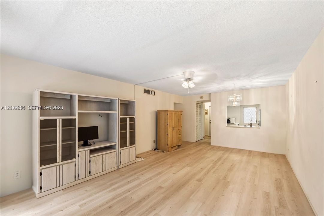 Living area featuring light-toned wood-finish flooring, built-in floor-to-ceiling cabinetry, recessed ceiling lighting, and a pass-through window to the kitchen