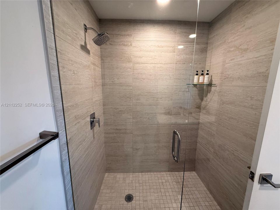 Walk-in shower featuring floor-to-ceiling stone-finish tile surround, frameless glass door, mosaic tile flooring, built-in glass shelving, and chrome hardware