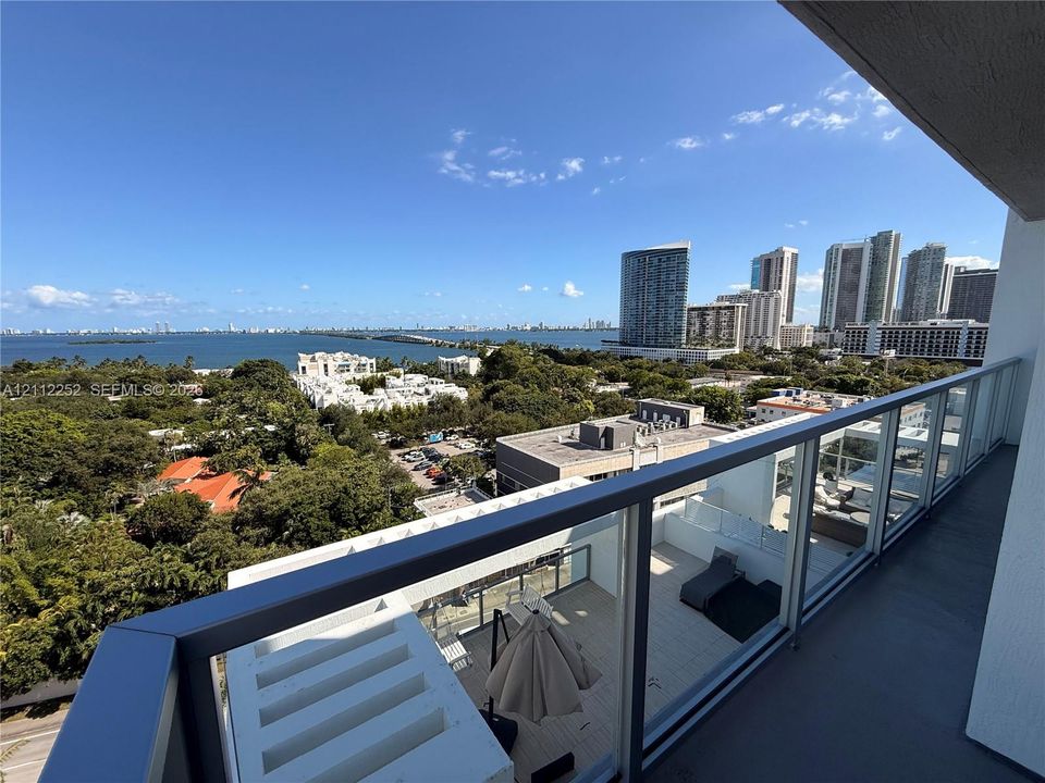 Covered balcony featuring glass railing panels and metal handrails, overlooking a coastal landscape with high-rise structures and open water