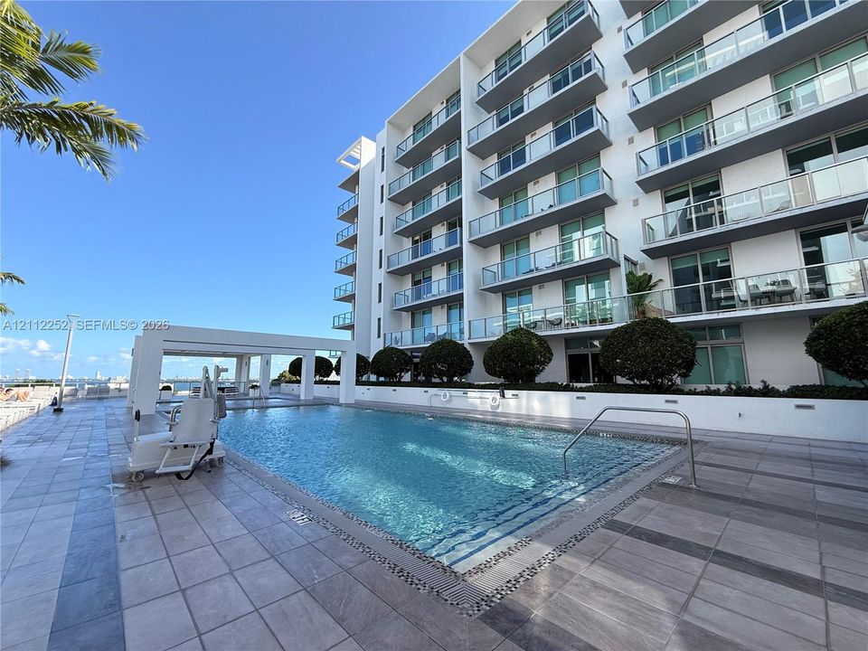 In-ground swimming pool with stone tiling, poolside cabana, paved terrace, and multi-story building exterior with glass-railed balconies