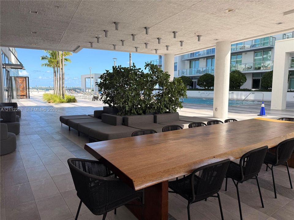 Covered outdoor terrace featuring large-format tile flooring, integrated ceiling lighting, structural columns, and direct access to a communal pool area