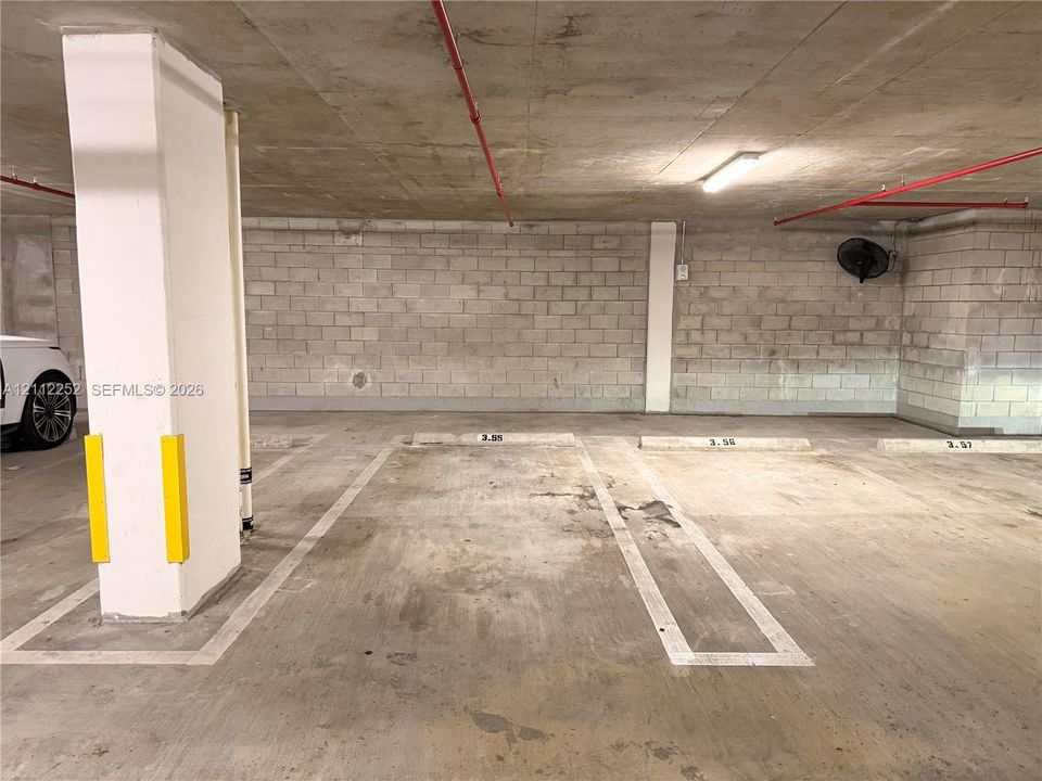 Covered parking area featuring concrete flooring, concrete block walls, painted support pillars with protective guards, and overhead lighting