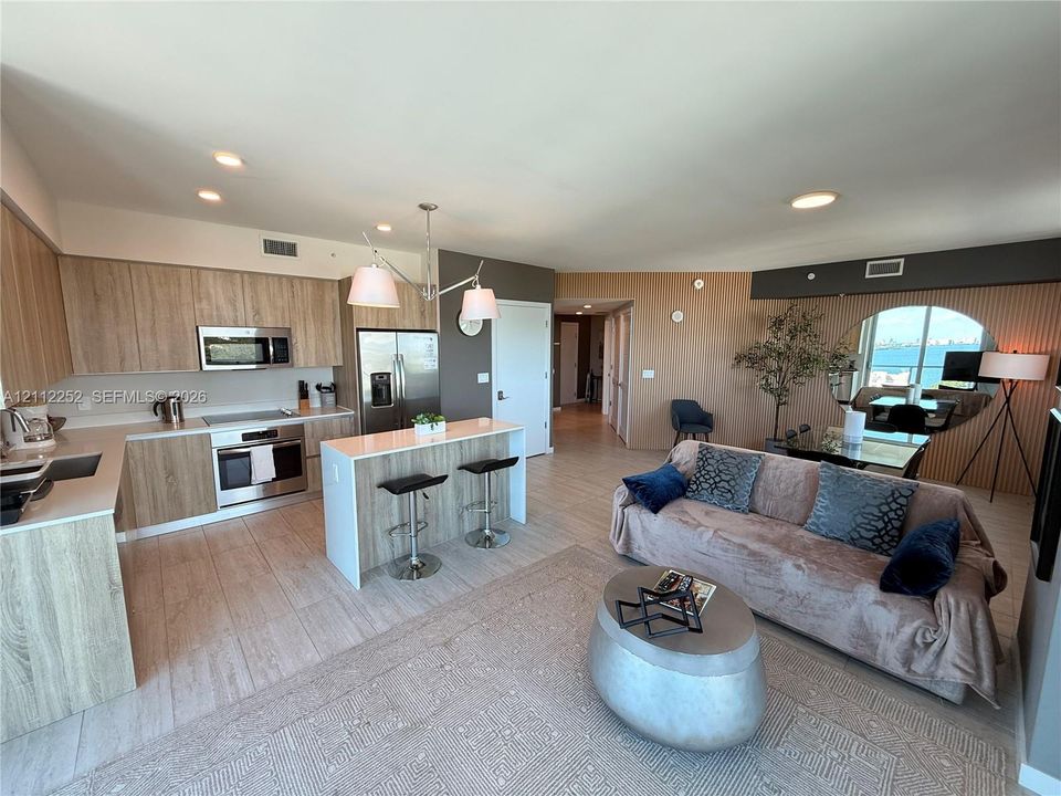 Open-concept kitchen featuring light wood-finish cabinetry, a central island with counter seating, stainless steel appliances, and wood-finish flooring throughout