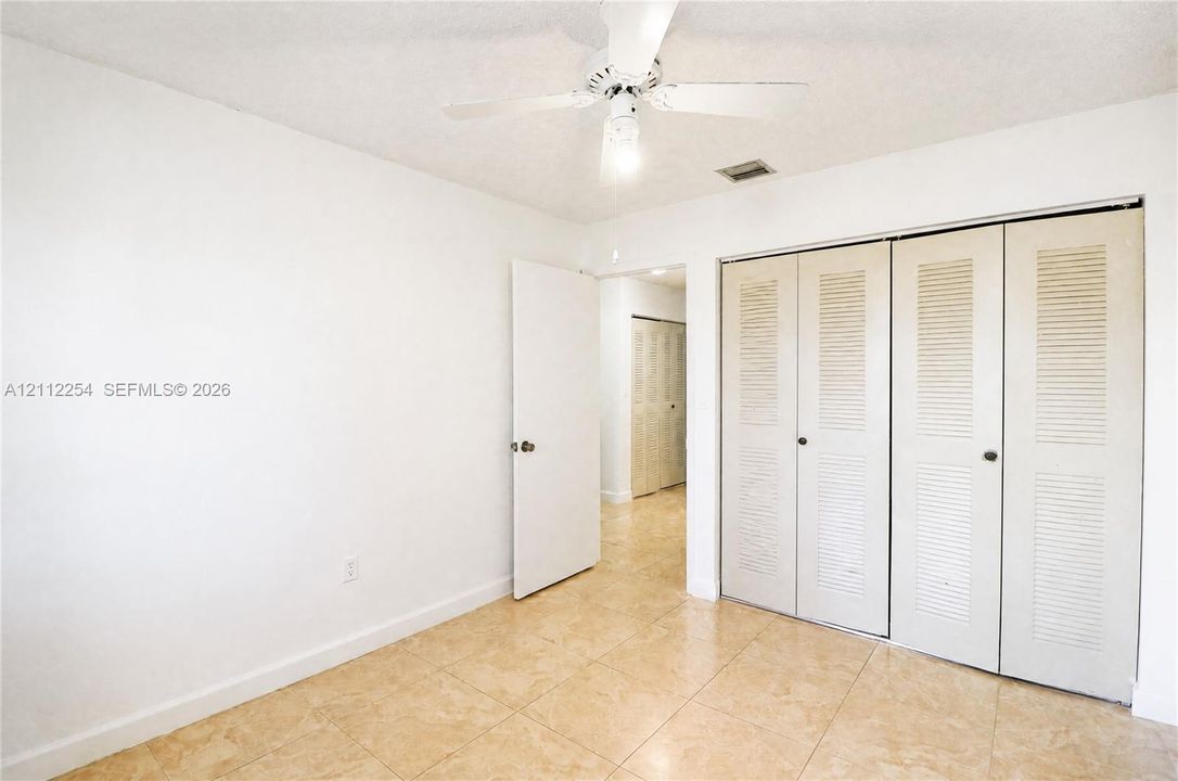 Neutral interior space featuring polished tile flooring, a multi-blade ceiling fan with integrated light fixture, and floor-to-ceiling double-door closets with louvered panels