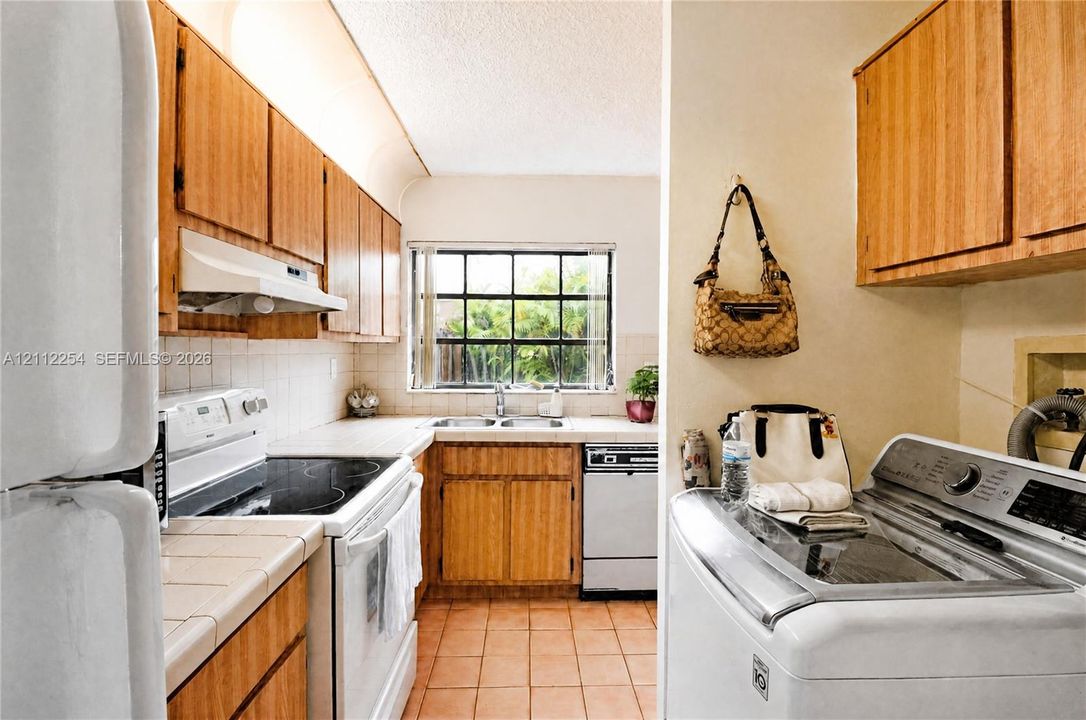 Kitchen featuring wood cabinetry, tile countertops with a coordinating backsplash, integrated range hood, and double-basin sink, accompanied by a dedicated laundry alcove with built-in cabinetry