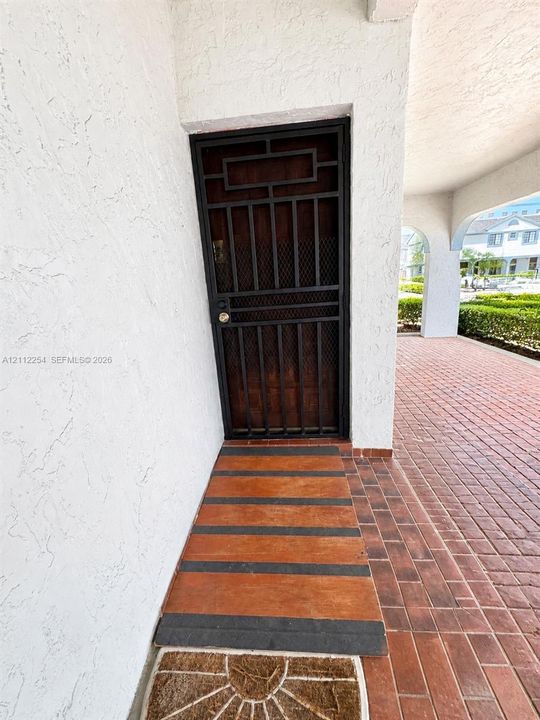 Covered entryway featuring a metal security screen door, stucco exterior walls, and red brick paver flooring