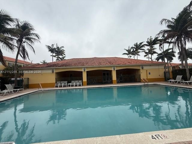 Swimming pool with a stone coping edge, surrounded by a paved deck, featuring a covered pavilion with arched architecture, a clay tile roof, and mature palm trees