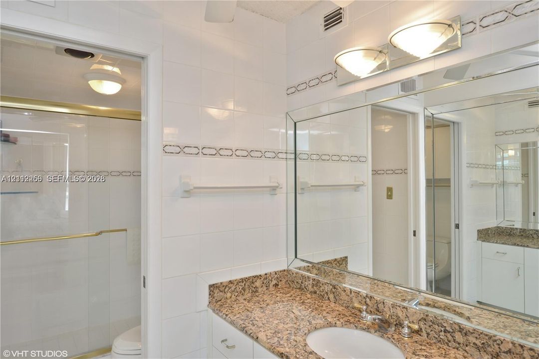 Full-width vanity with stone-finish countertop, undermount sink, integrated mirror, glass-enclosed shower with gold-tone hardware, and white wall tiles with decorative accent trim