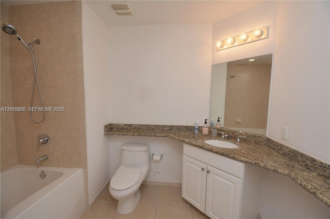 Bathroom featuring a stone-surface vanity with an integrated oval sink, white cabinetry, a full-width vanity mirror, a tiled shower-tub combination with a handheld showerhead, and a neutral-toned tile floor