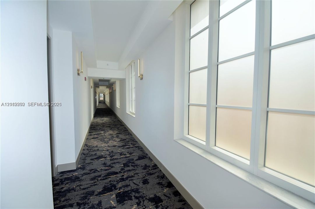Hallway featuring expansive windows, patterned wall-to-wall carpeting, and wall-mounted light fixtures