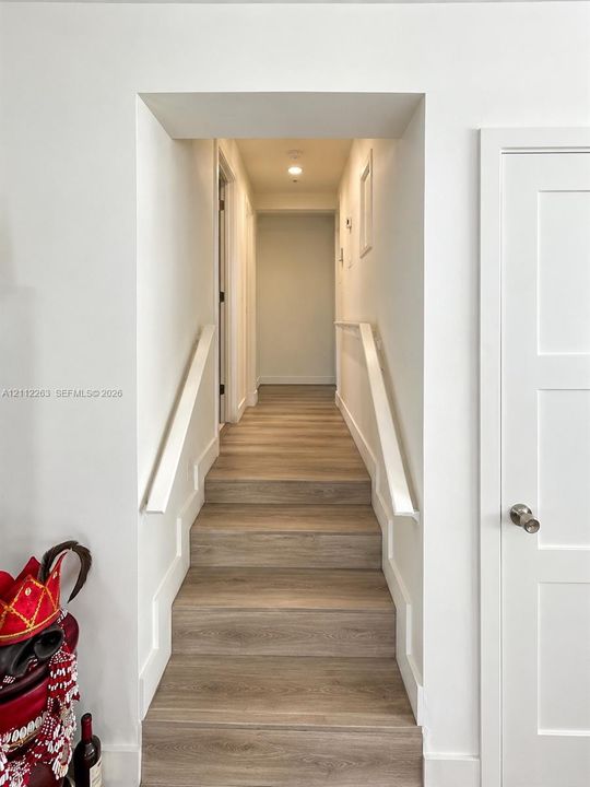 Staircase featuring wood-finish flooring, crisp white trim, and white handrails, complemented by a solid core shaker-style door with a brushed nickel handle