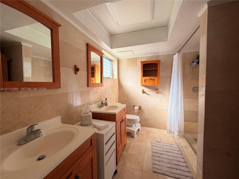Dual vanity with wood-finish cabinetry, stone-tiled wall surfaces, integrated lighting tray ceiling, and a walk-in shower enclosure