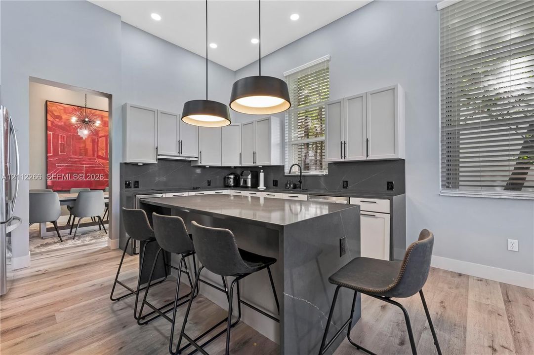 Kitchen featuring a central island with a solid-surface waterfall countertop, white cabinetry with dark hardware, dark solid-surface backsplash, wood-finish flooring, and dual pendant lighting